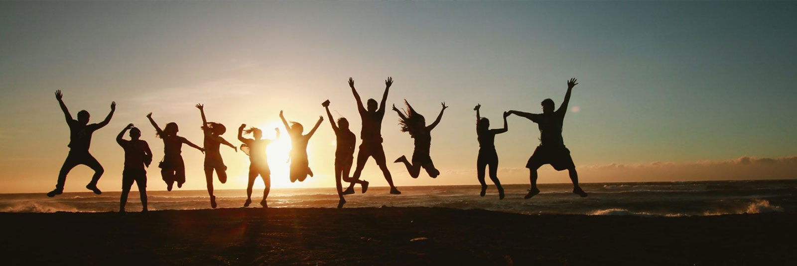 Während am Strand die Sonne untergeht, springt eine Gruppe von Menschen fröhlich in die Luft